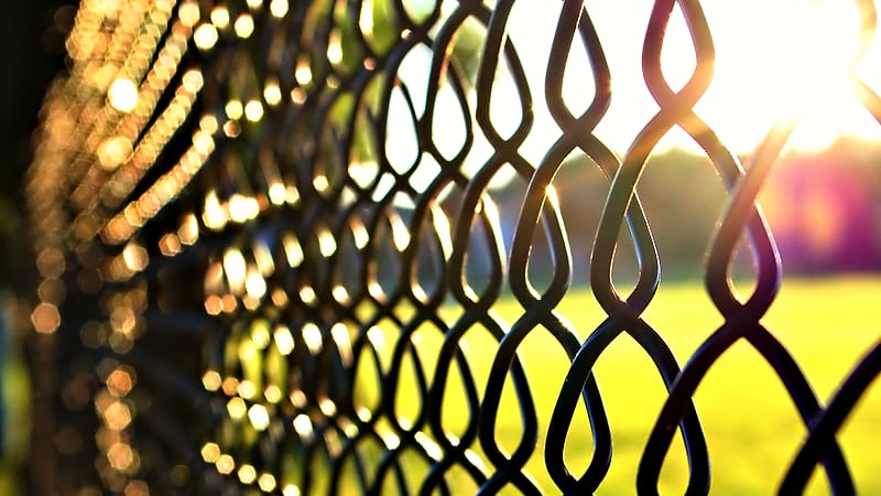 A tall chain link fence topped with coiled wire against a flat overcast sky above a paved empty yard