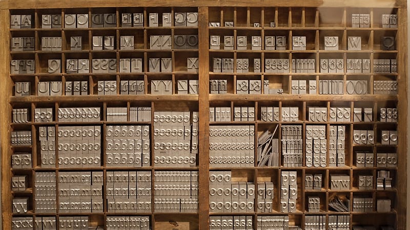 A gray archive box with its lid removed on a metal shelf, holding upright folders with handwritten paper tabs