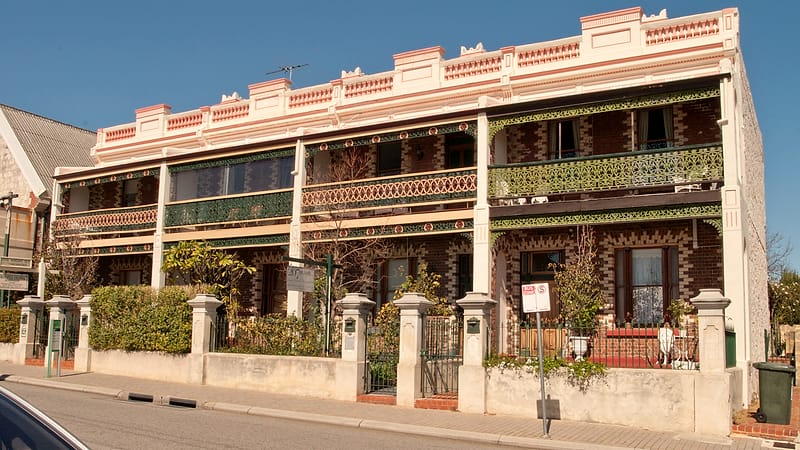 A narrow residential street of attached townhouses with bay windows, stone steps and parked cars under flat afternoon light