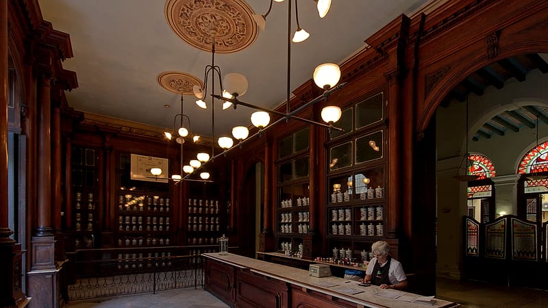 Rows of white labeled bottles and cartons on metal shelving behind a pharmacy counter under even lighting