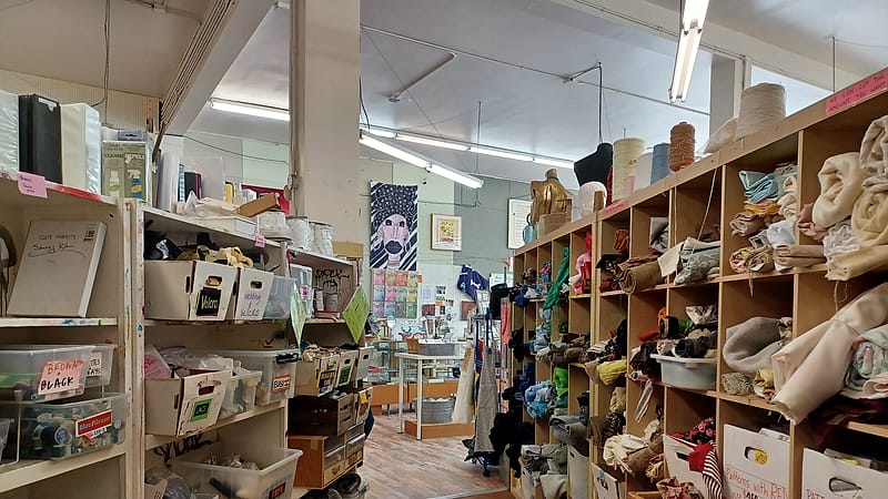 A crowded second-hand shop interior with mixed goods stacked on shelves and a rail of items in the foreground