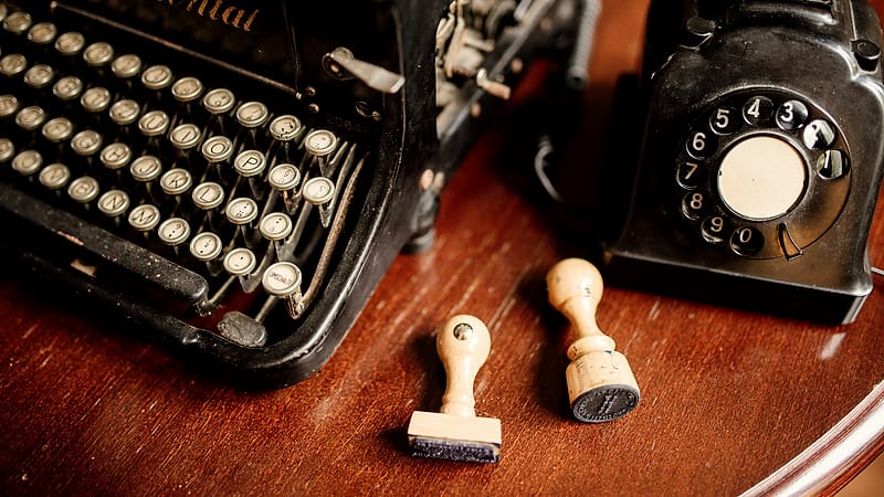 A plain black desk telephone with a coiled cord sitting beside a closed notebook on a bare office table in daylight