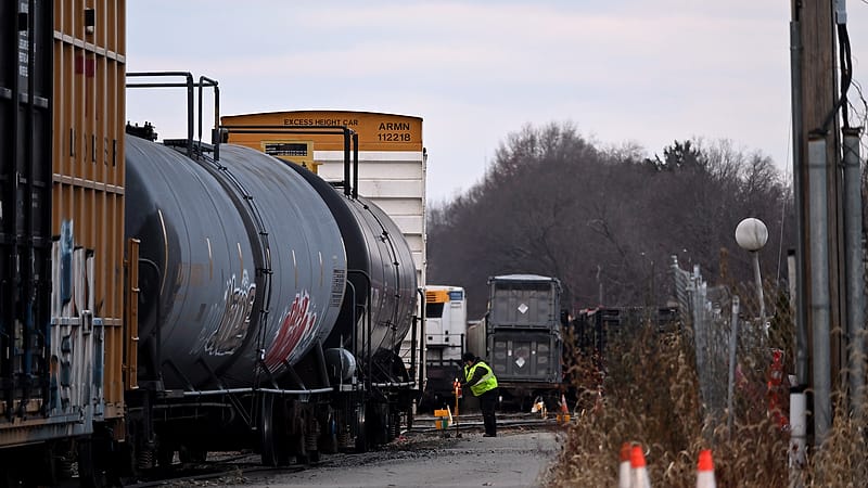 A railroad worker in a high visibility vest and hard hat standing beside freight cars in a rail yard at dusk