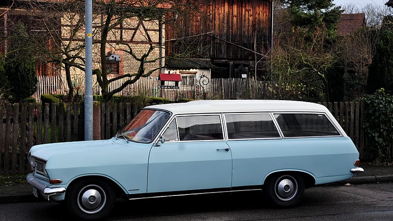 A dusty sedan with flat tires parked at the edge of a gravel lot beside a fence, weeds growing around the wheels
