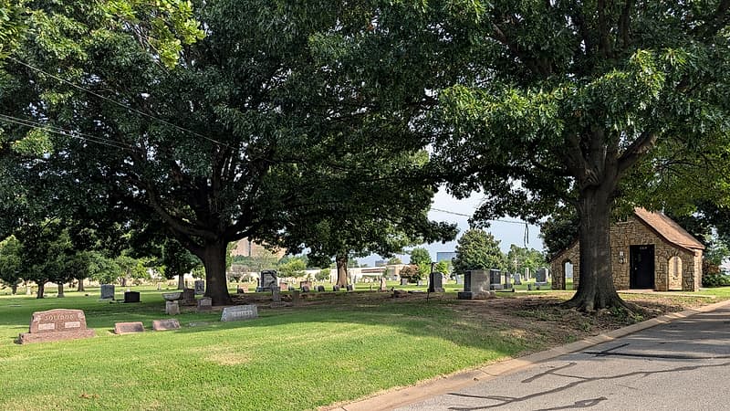 A mown cemetery lawn with flat bronze markers set flush with the grass and a line of mature trees along the far edge