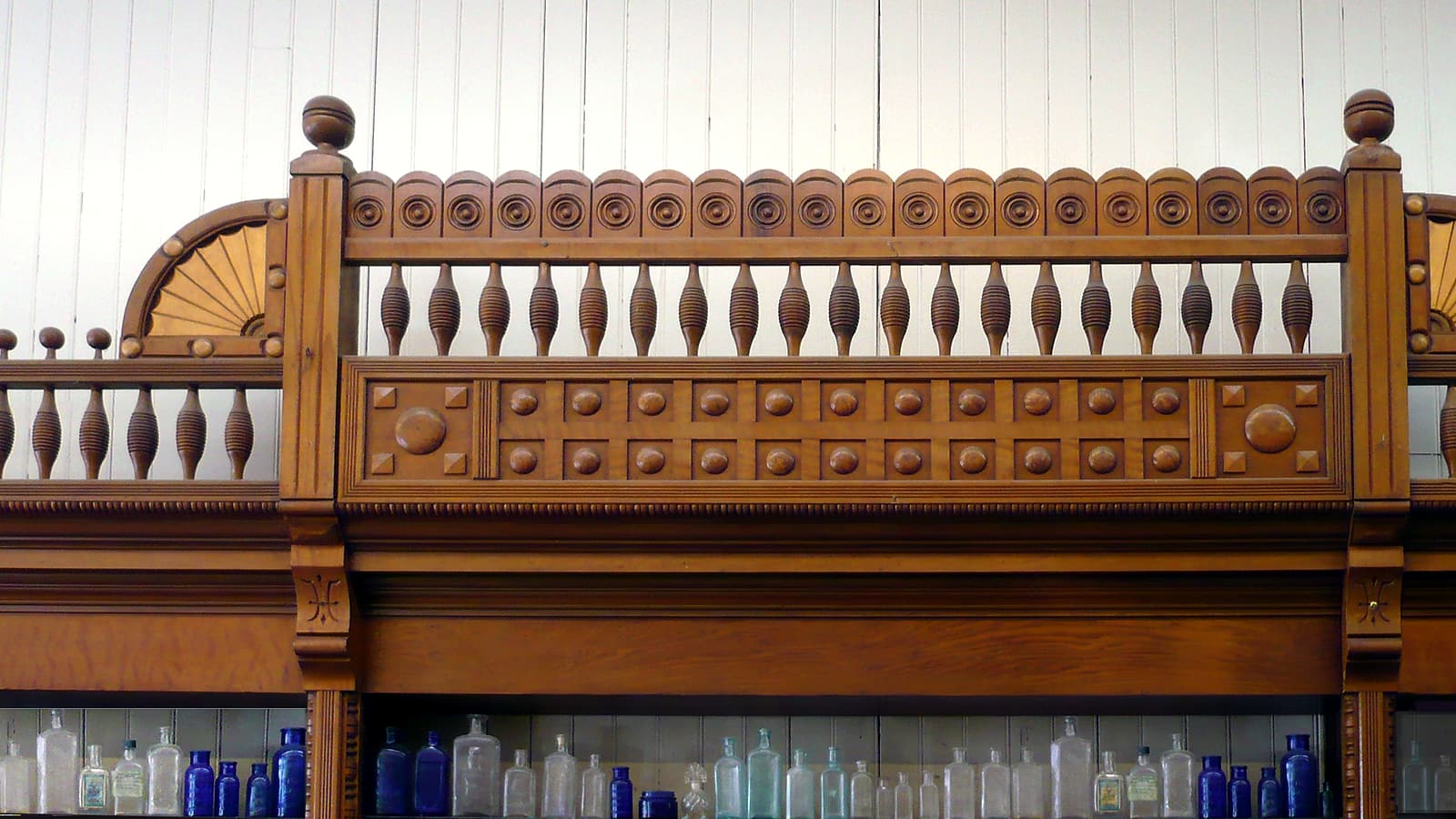 A pharmacist in a white coat standing at a counter reviewing a document, seen from behind against shelves of stock