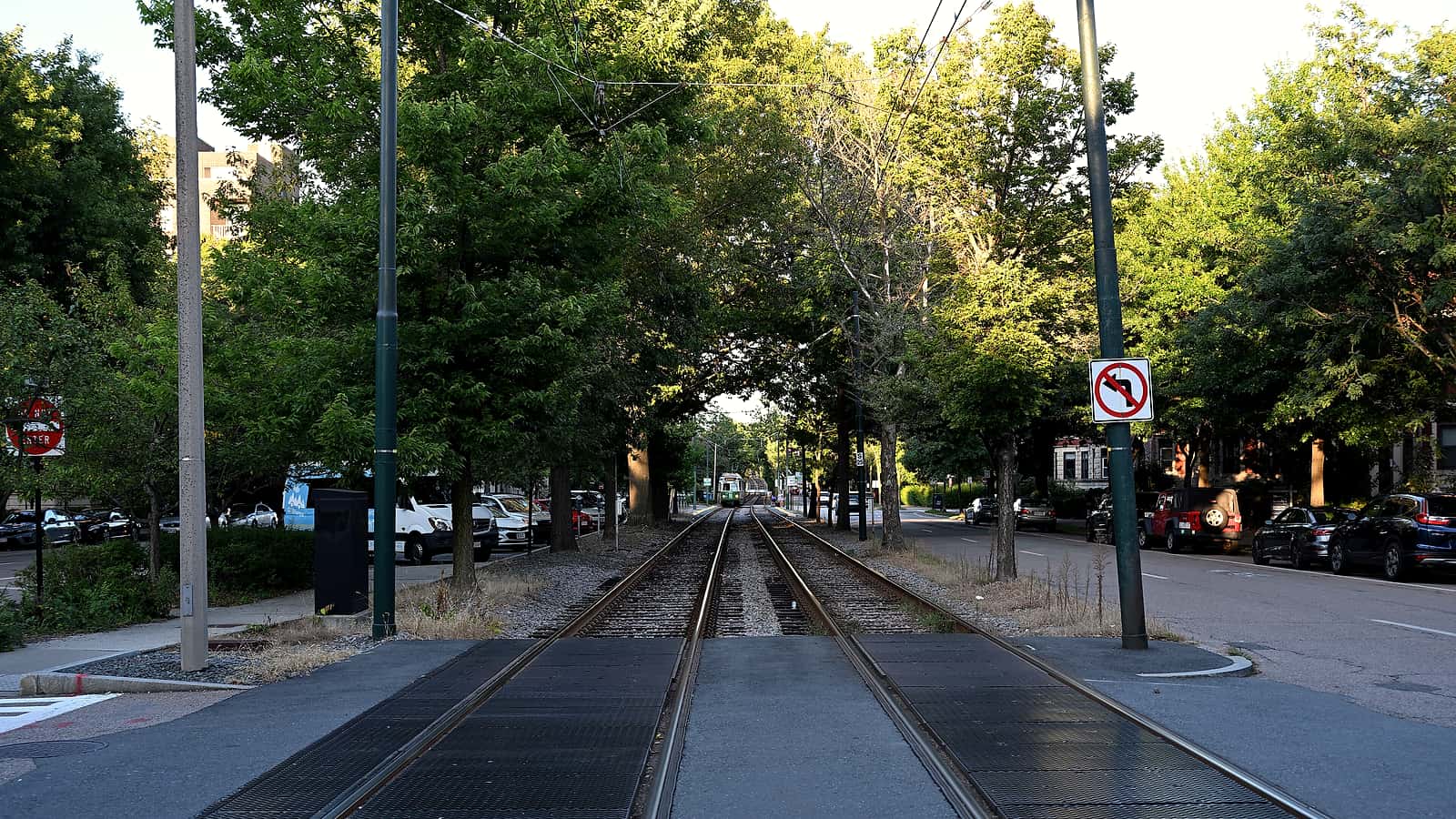 A rural road approaching a single track crossing with tall grass and brush growing close to the rail line