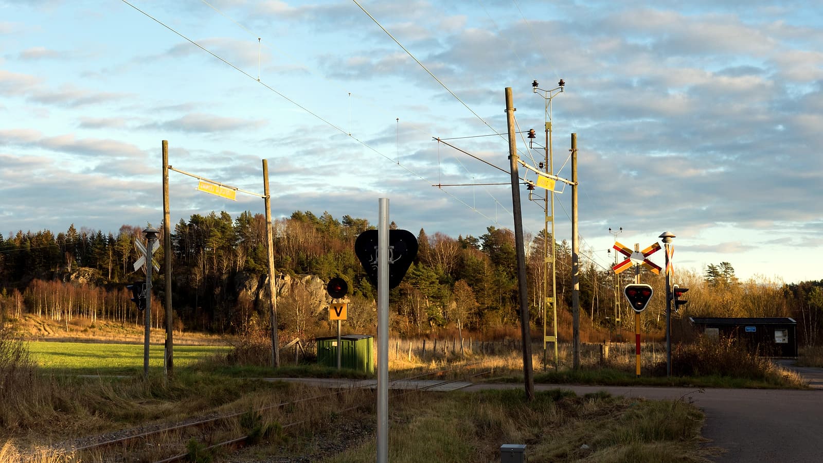 A red and white crossing gate lowered across a two-lane road beside flashing signal masts and a signal bungalow
